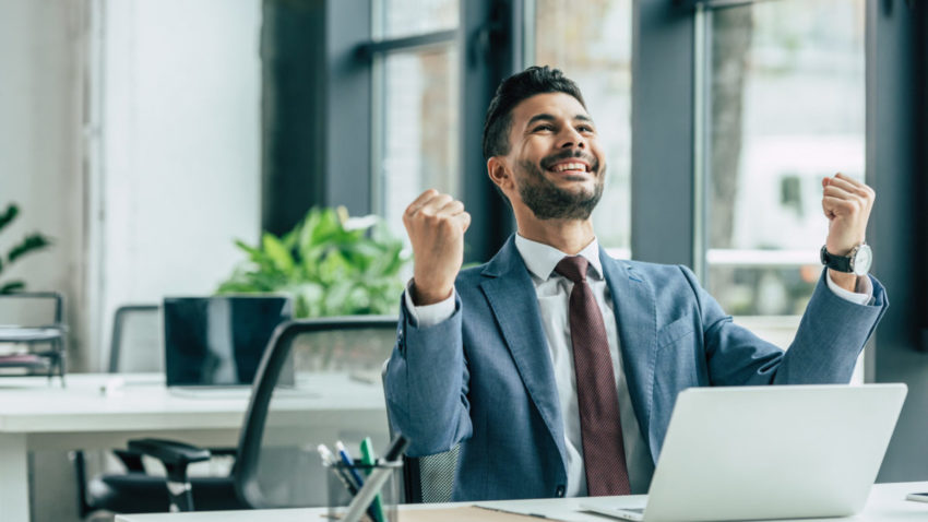 happy businessman looking up and showing winner gesture while sitting at workplace
